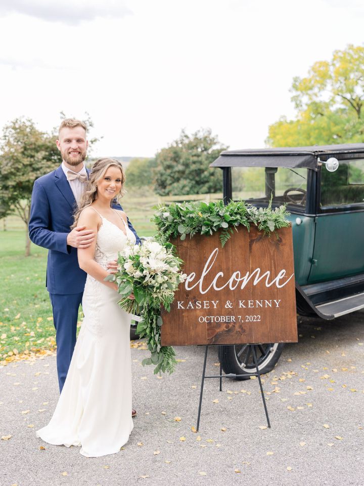 Bride and Groom pose in front of their wooden welcome sign and vintage car holding their white and green wedding flowers.