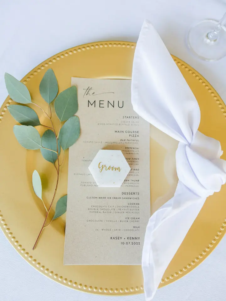 Gold charger with a white napkin, tied in a knot, a kraft menu and a piece of eucalyptus