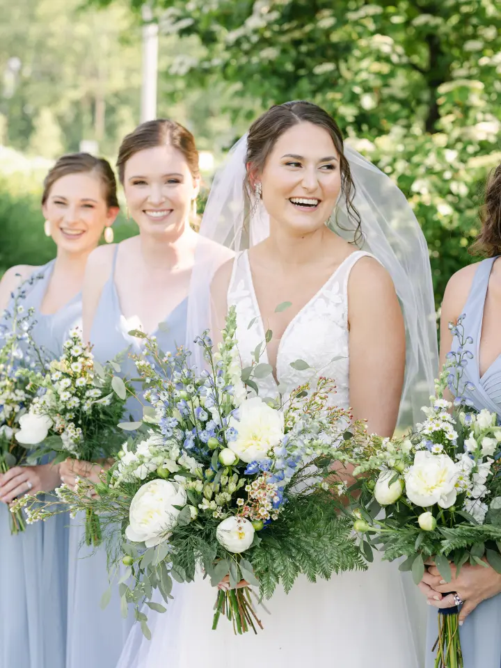 Bride laughing and looking at her bridesmaids, all wearing light blue chiffon gowns carrying blue white and green bouquets