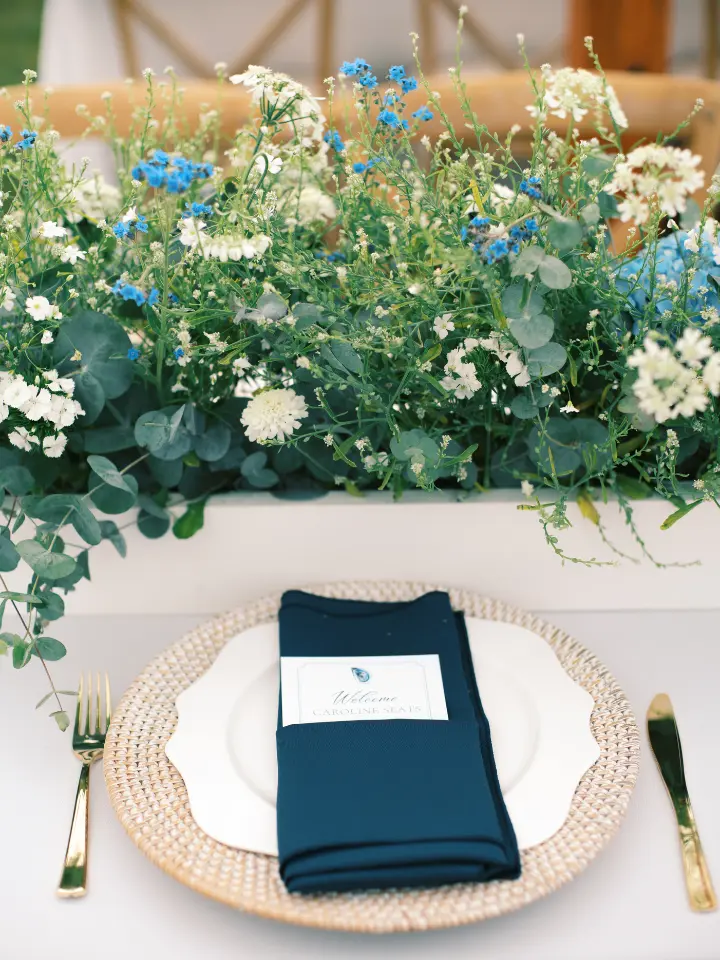 Up close table setting with greenery box runner, navy napkin on top of a white plate with a textured brown charger