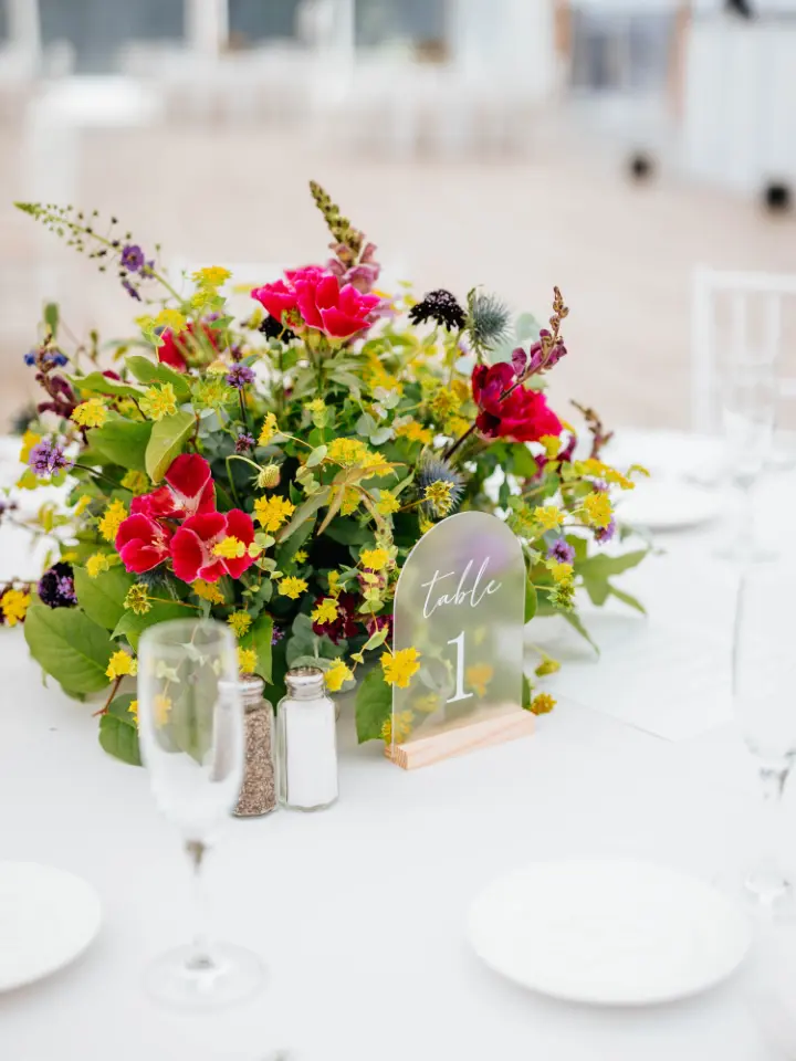Round small table centerpice on a white linen with mostly greenery, yellow and pink accents