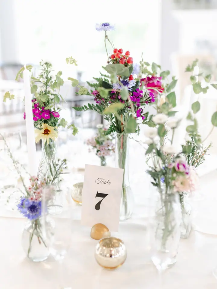 Bud vases on a round table with wildflowers and a simple white table number with the number "7" on it against a white linen, up close