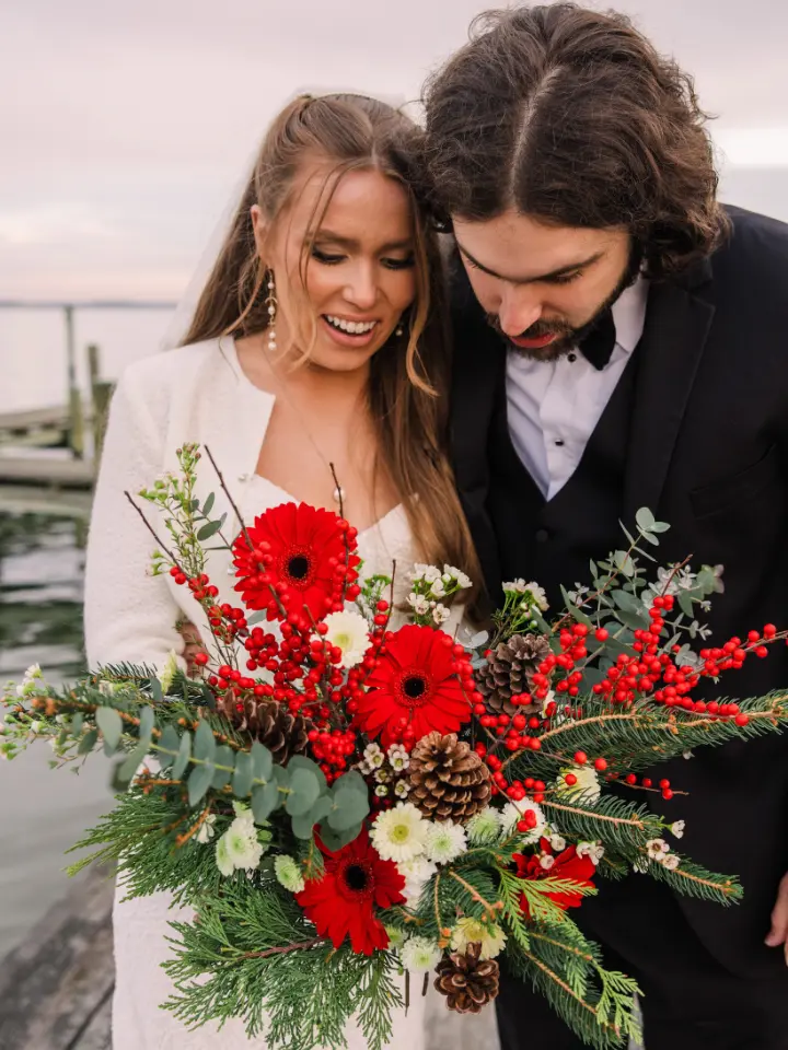 Bride and Groom smiling and looking down at a large bouquet of red ,white and green