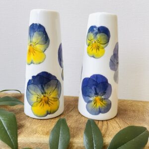 Two pressed flower bud vases with pansies sit on a wooden shelf with greenery.
