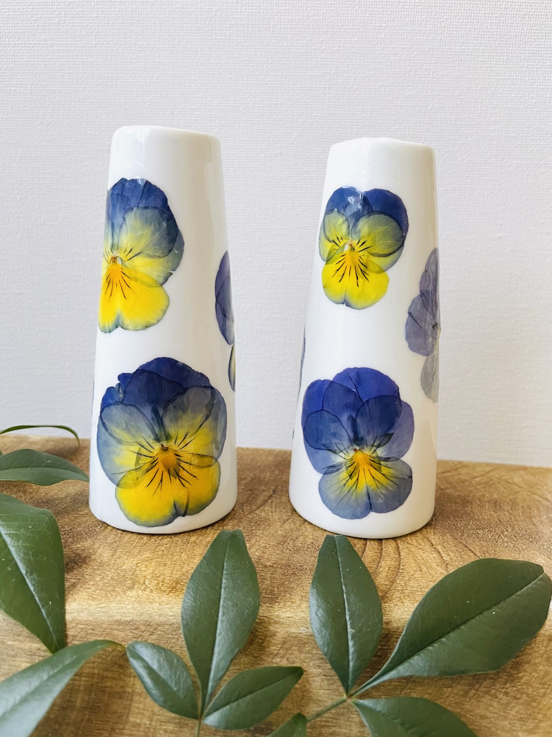 Two pressed flower bud vases with pansies sit on a wooden shelf with greenery.