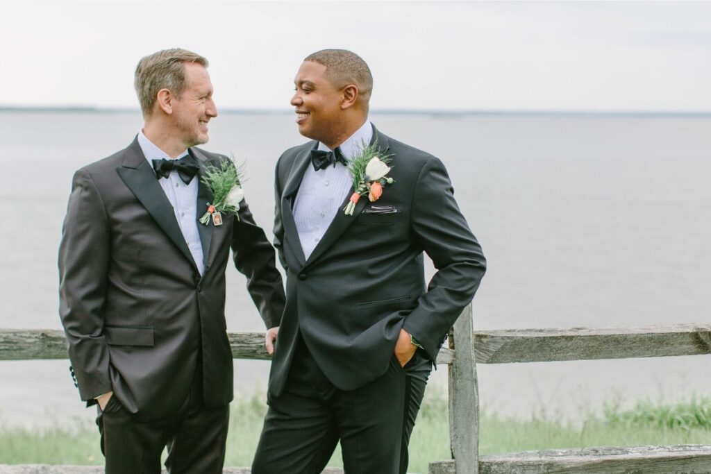 Grooms stand at a wooden fence looking towards each other while smiling.