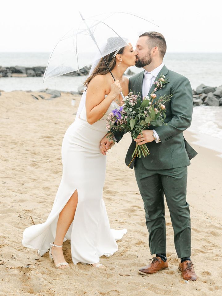 Bride and groom kiss on the beach under a clear umbrella.