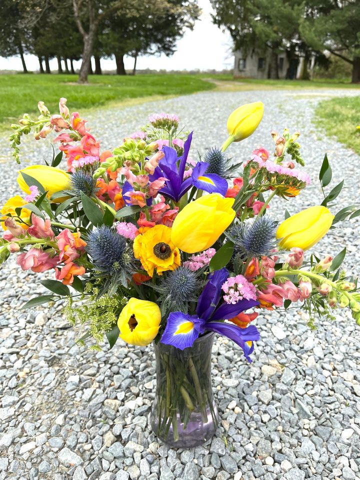 A bright and colorful Mother's Day Vase flower arrangement sits outdoors on a gravel driveway.