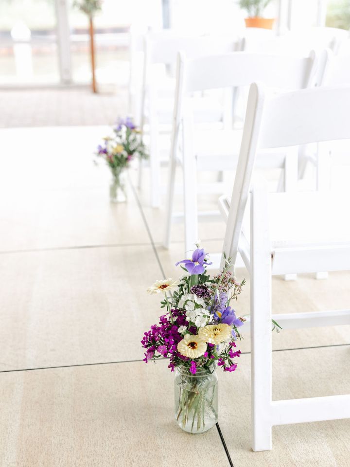 Mason jars filled with pastel spring flowers line the aisle for a wedding ceremony.