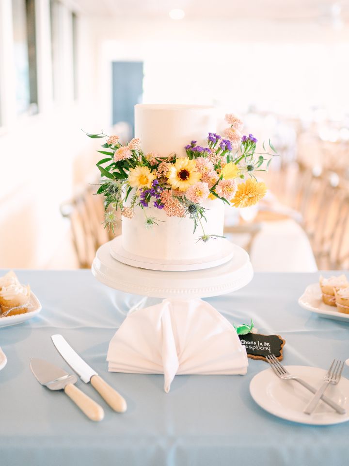 A 3 tier white wedding cake with spring flowers on it, on a blue table cloth.
