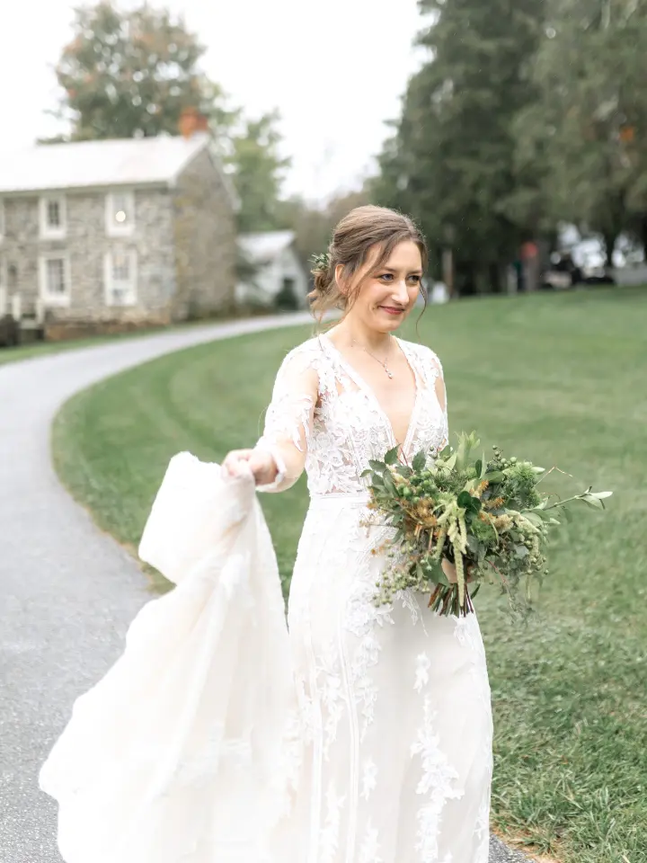Bride holding the edge of her dress and a full greenery bouquet walking out of camera