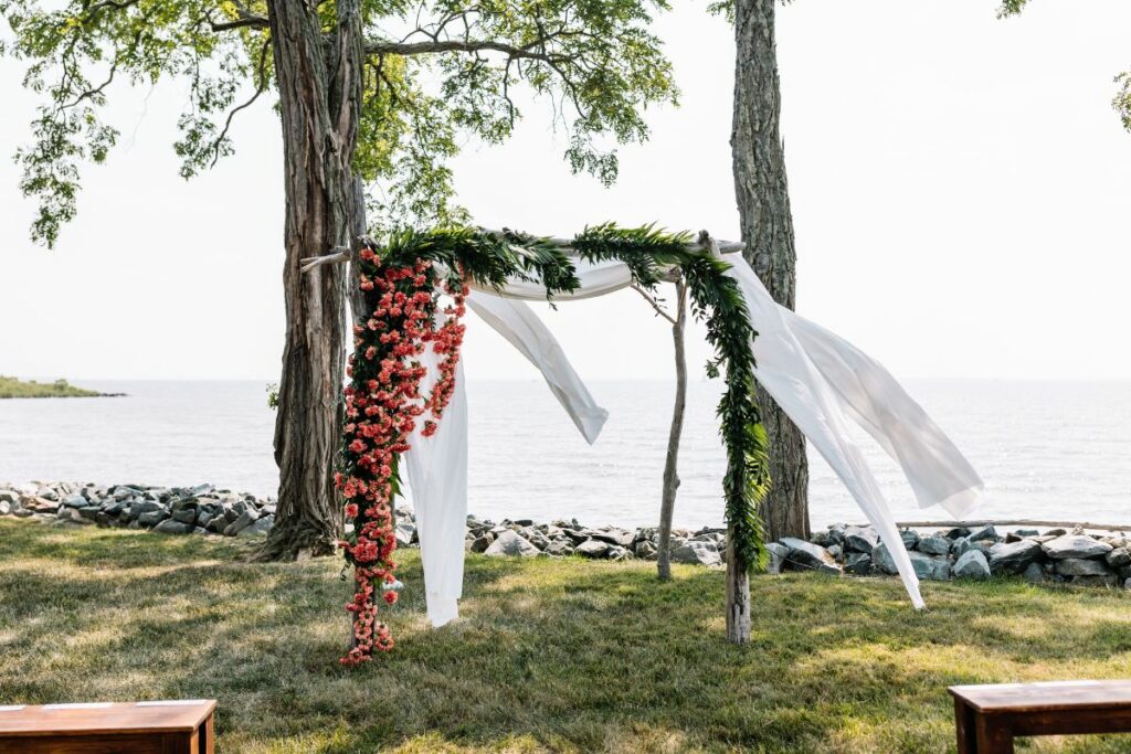 View of a rustic chuppah with white fabric, and greenery and orange floral curtain.