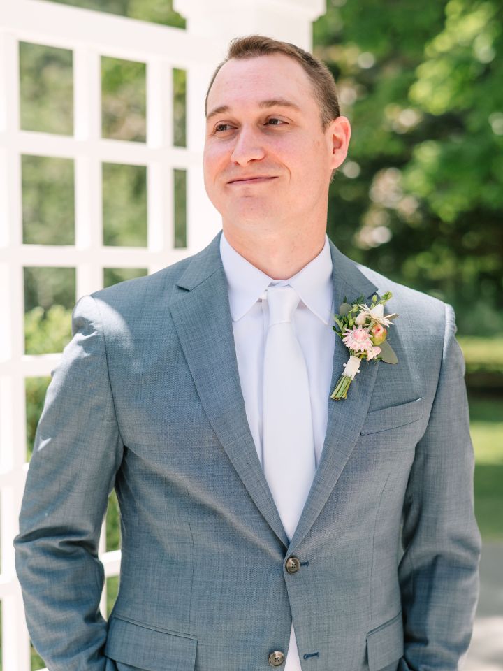 Groom in a grey suit wears a pink and white boutonniere under a white pergola.