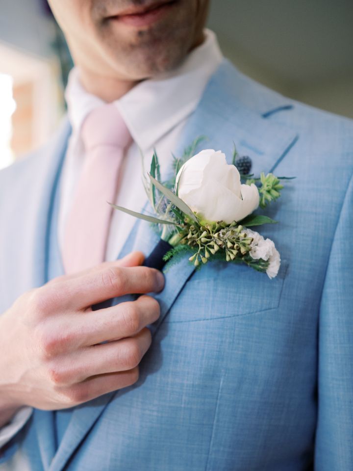 Groom in a pale blue suit with light pink tie attaches his white peony boutonniere.