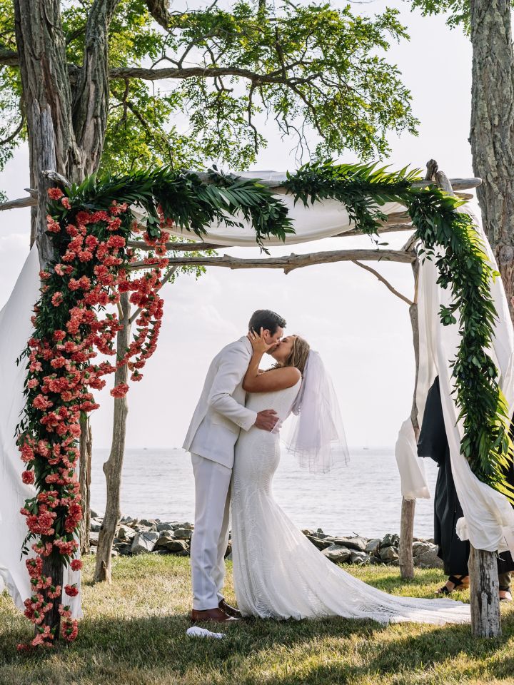 Bride and groom kiss under a chuppah with greenery and orange flowers.