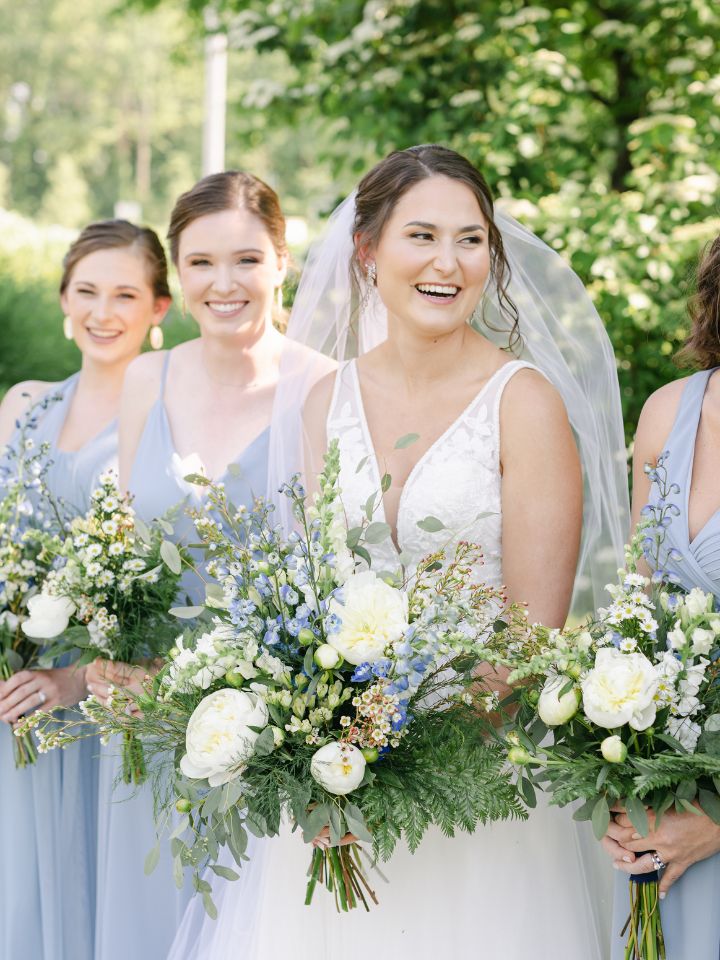 Bride smiles at her bridesmaids holding blue and white wedding flowers.