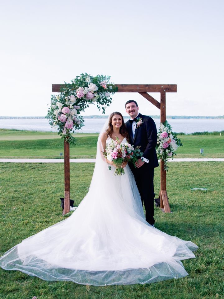 Bride and Groom stand under a wooden arch with pink flowers on it.