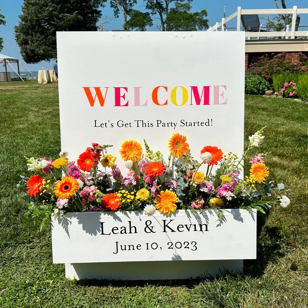 A welcome flower box sign with orange and yellow accents at a wedding