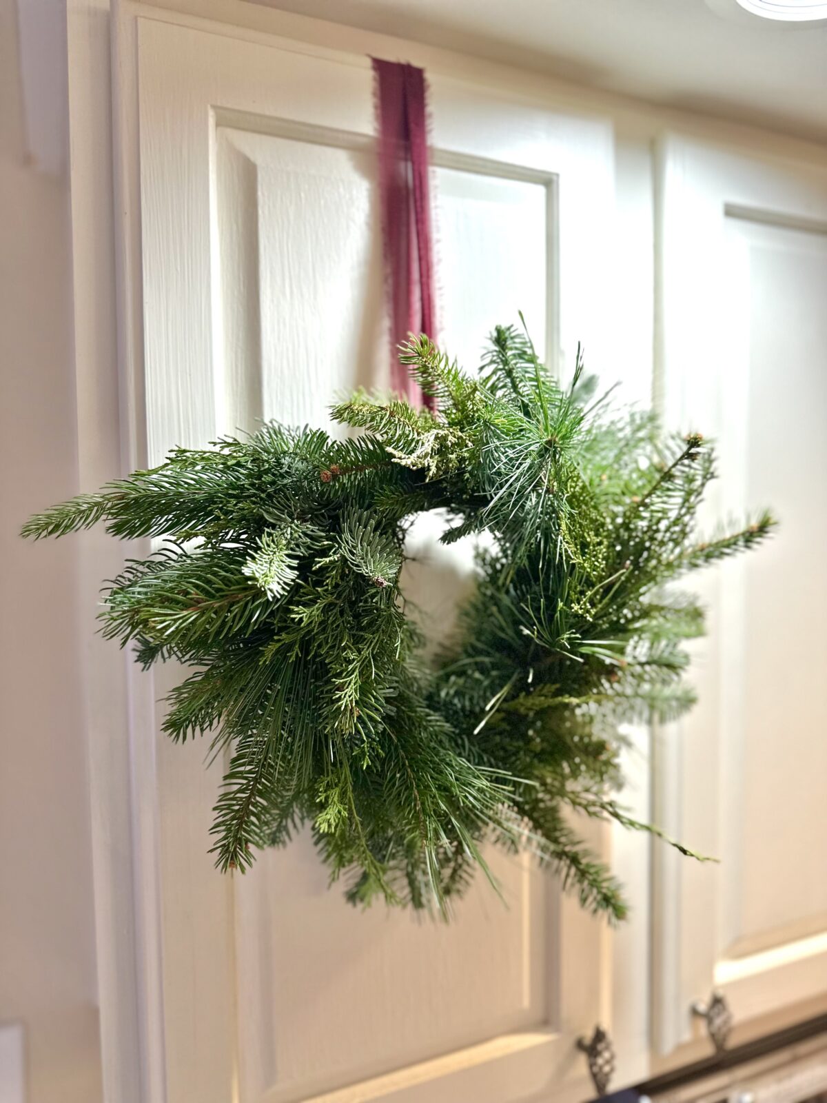 Mini greens wreath hangs from a burgundy ribbon on a white kitchen cabinet.