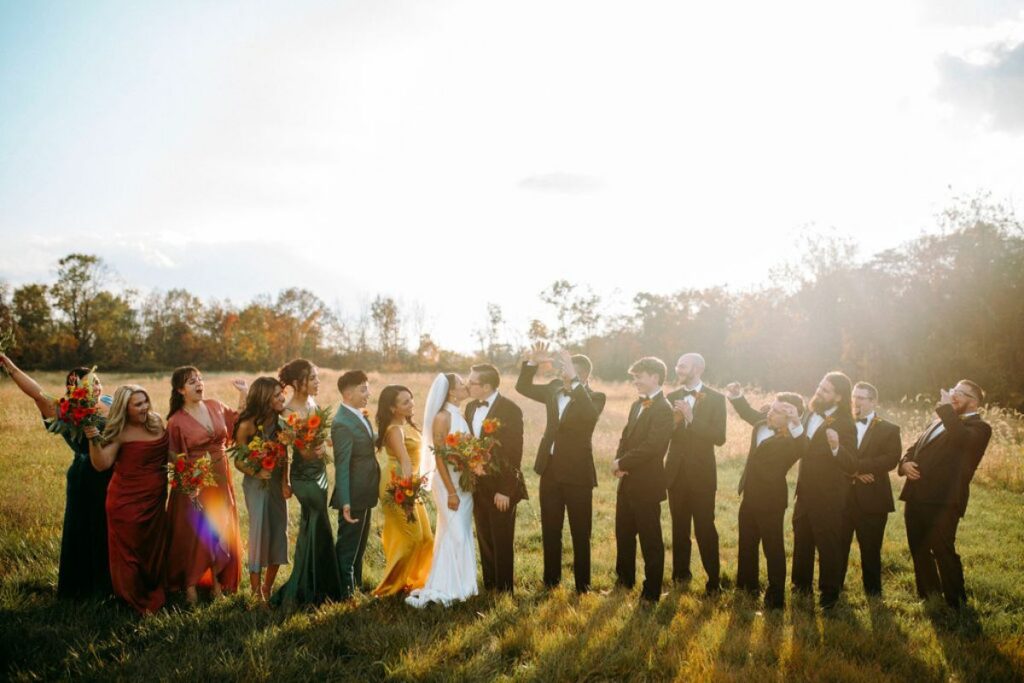 Bride and groom kiss surrounded by cheering bridal party in field at Hazelwood Weddings