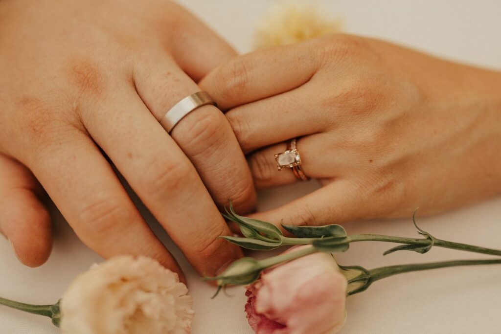 Bride and groom hold hands, showing off wedding rings surround by pastel peach and pink flowers