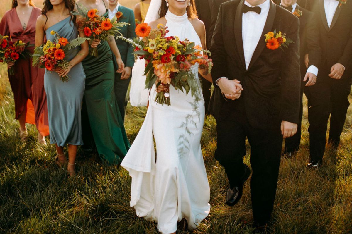 Bride and groom walk with bridal party behind them holding orange flower bouquets and wearing orange flower boutonnieres