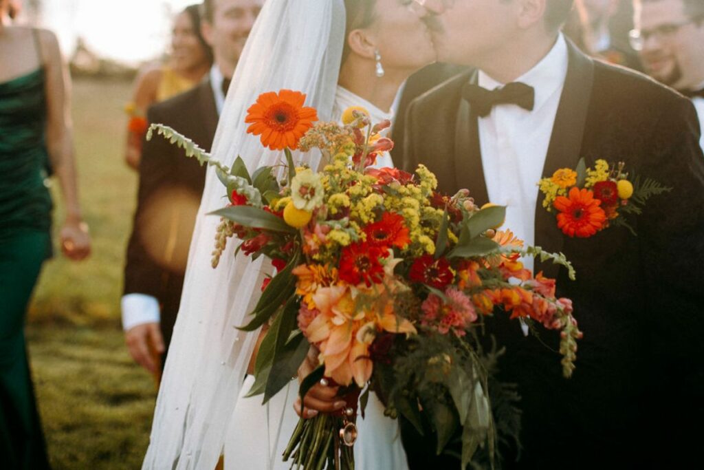 Bride holds a wedding bouquet with orange, peach, and yellow fall flowers to the camera while kissing groom