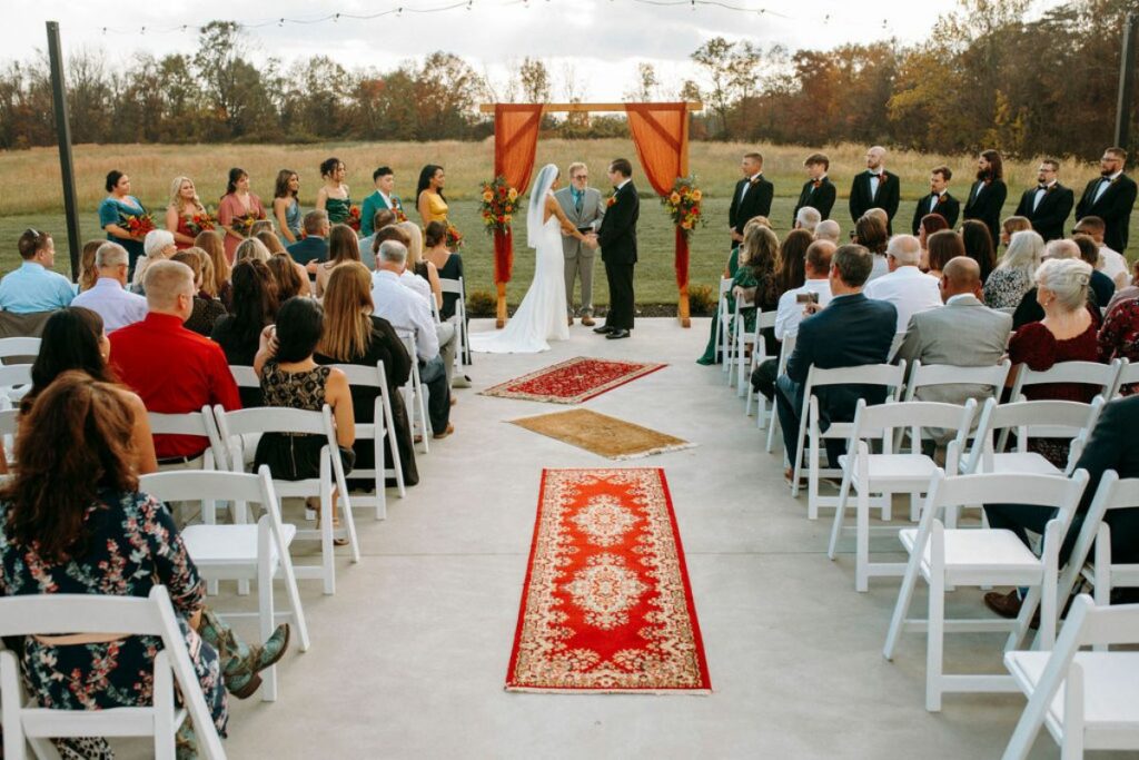 Bride and groom stand at end of wedding aisle underneath a rustic arch with orange chiffon fabric and autumn flowers. There are orange tone rugs on the aisle walkway and guests sitting in white chairs