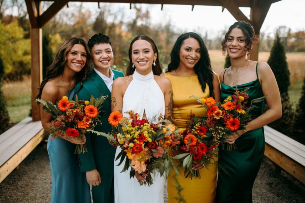 Bride is surrounded by bridal party wearing jewel tone outfits and holding autumnal bouquets