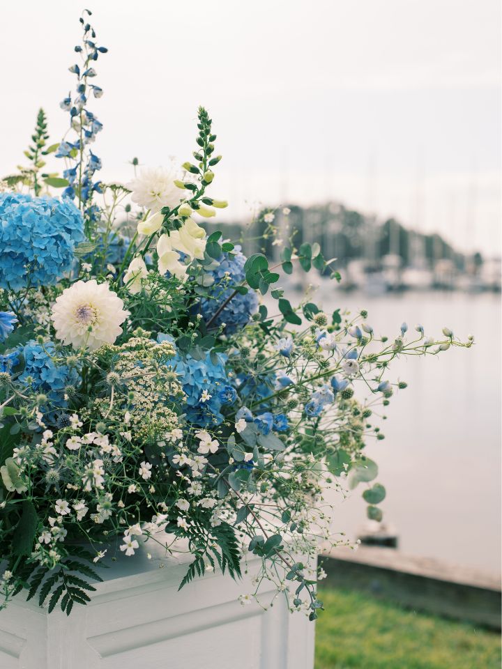 Blue and white flower arrangement on a white pedestal in front of the water for waterfront wedding ceremony