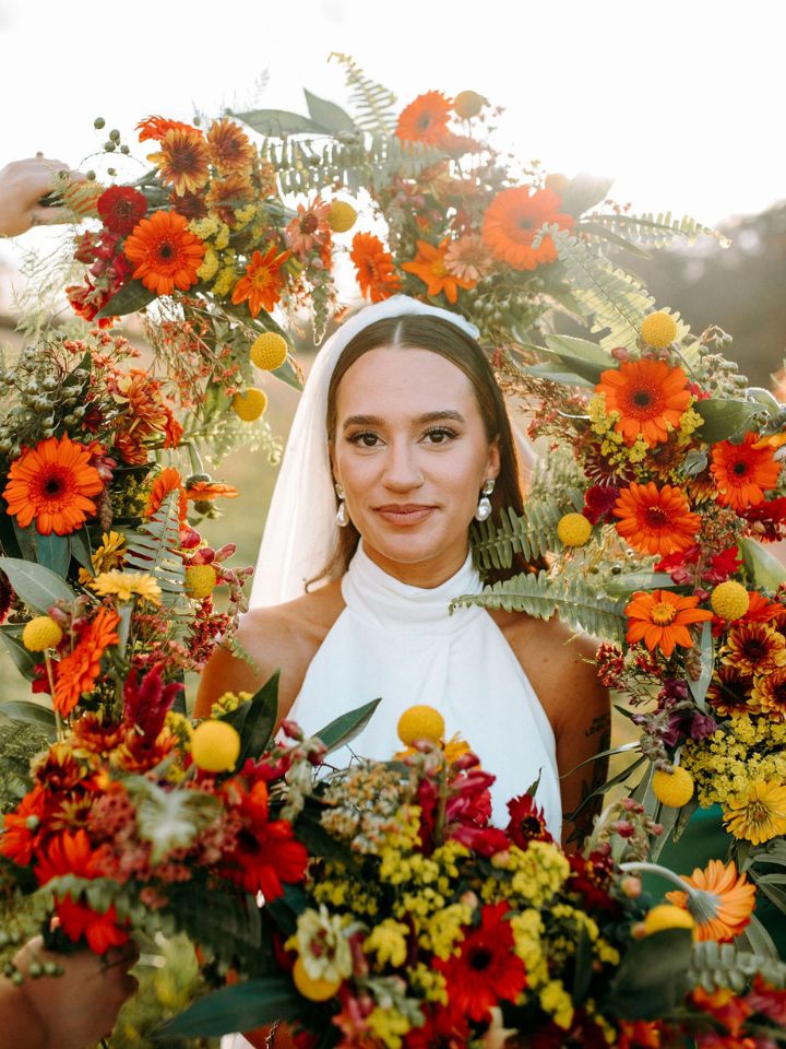 Bride in high neck retro style wedding dress surrounded by bridesmaids' bouquets of orange and yellow flowers and greenery