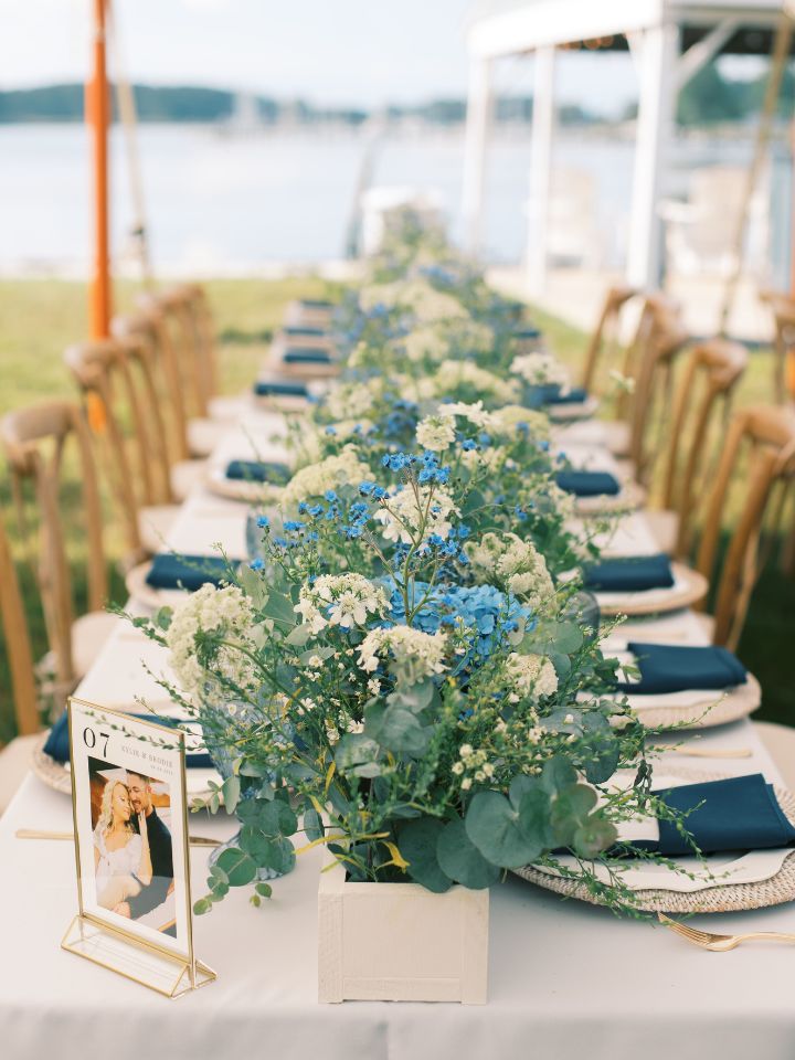 Long wedding reception table with white planter box centerpiece filled withg greenery, blue and white flowers. 