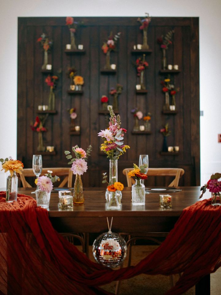 Bride & groom's head table at retro vibes wedding has bud vases on table and a dark wood wall behind the table with shelves on it that hold bud vases and square votive candles