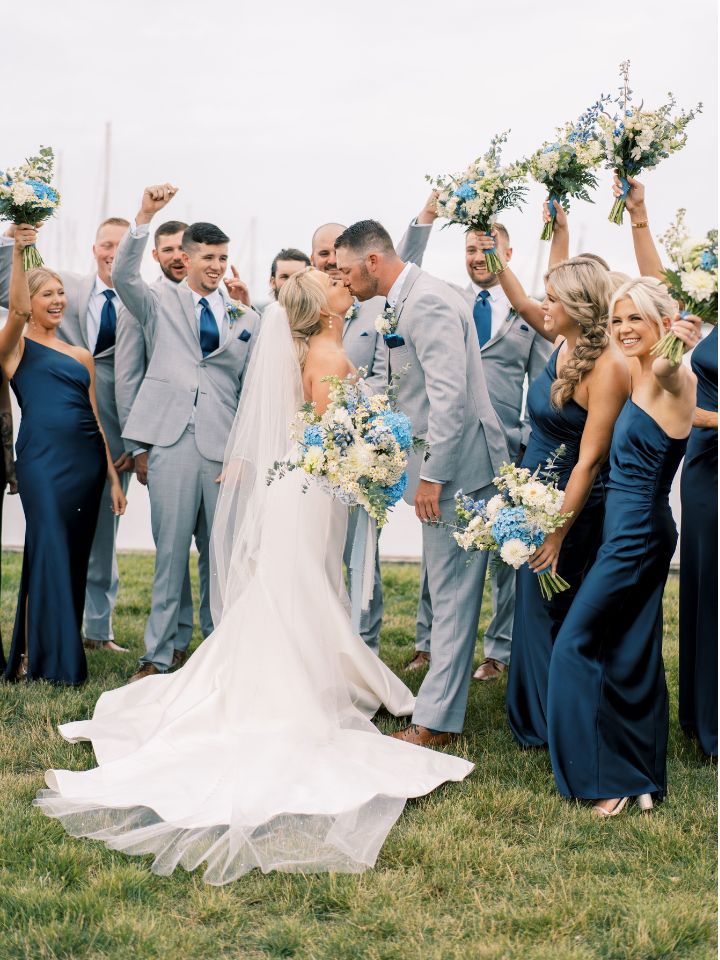 Bridal party cheering for bride and groom. Bouquets with blue flowers held in air by bridesmaids in blue dresses and groomsmen in grey suits