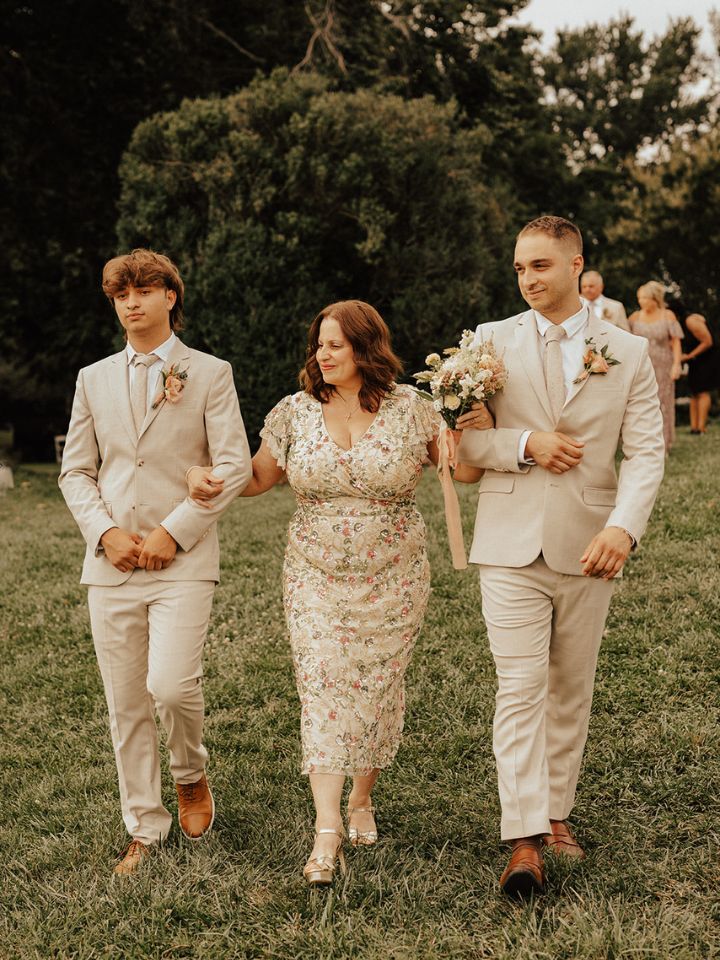 Mother of the bride carries a small bouquet and walks with two ushers in neutral suits