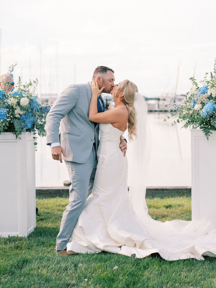 Bride and groom kiss at the end of the wedding aisle in front of the waterfront and pedestal flowers