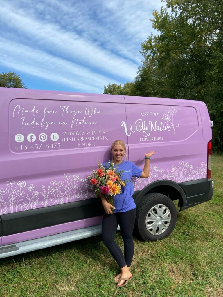 Lizzy stands in front of the purple van with a bouquet of flowers.