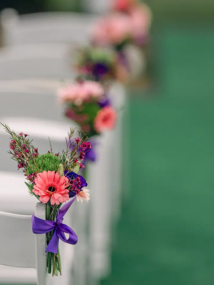 pink and purple flower bundles with greenery tied with purple ribbon to white chairs