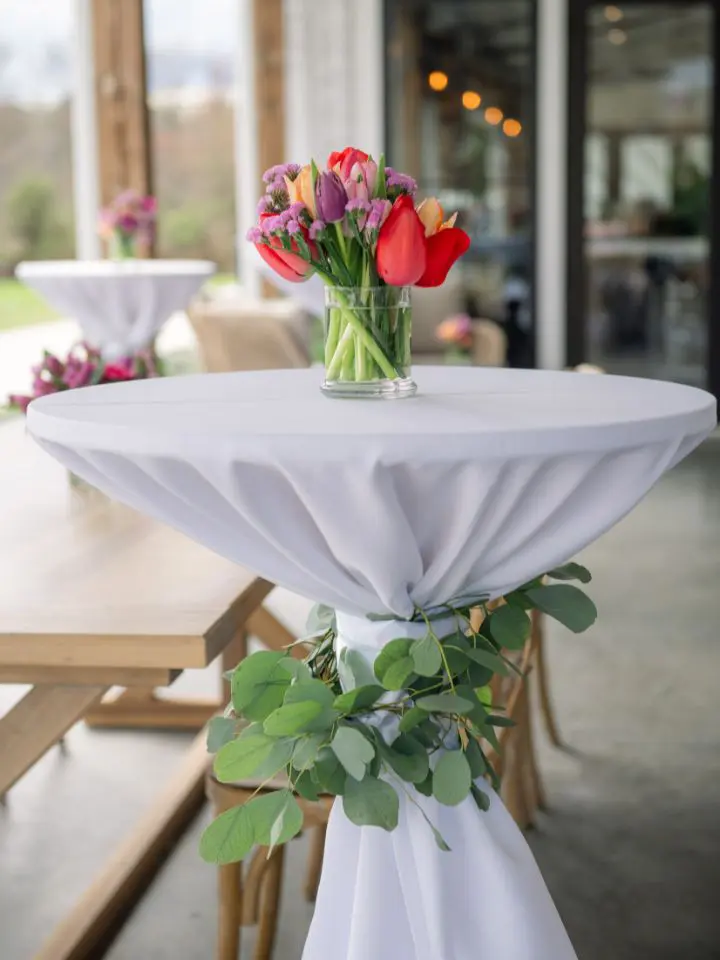 Hightop cocktail tables with white linens, eucalyptus garland wrapped around, and small bud vases filled with colorful flowers