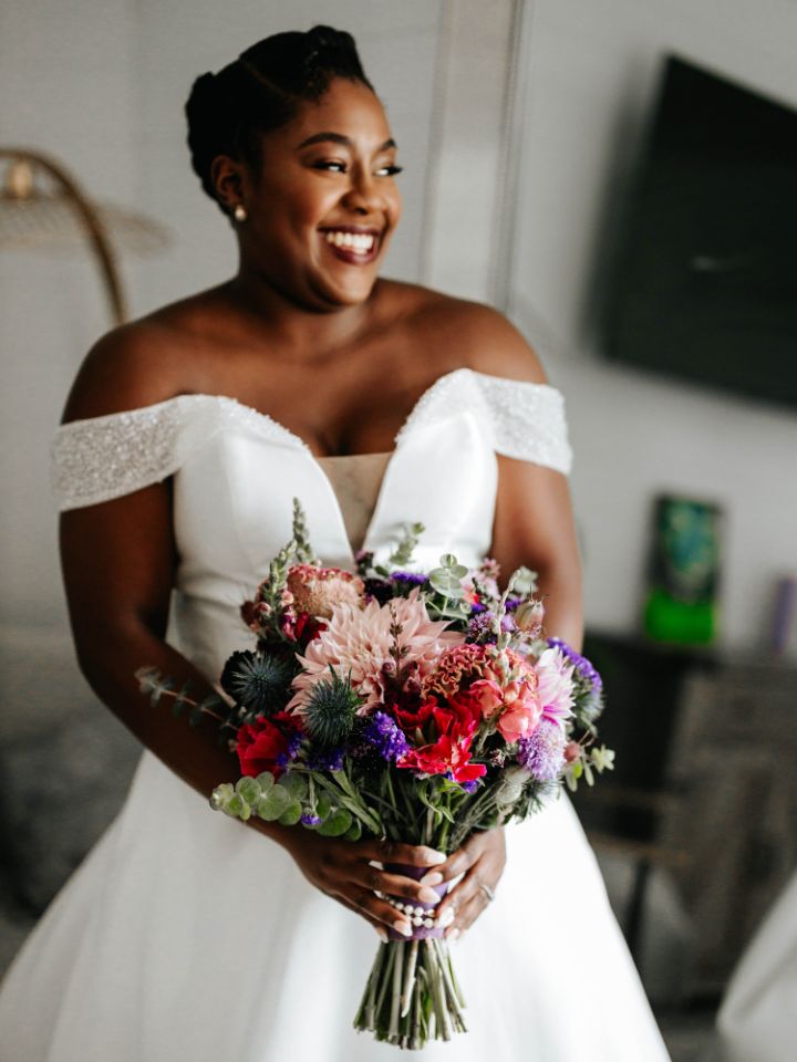 Bride smiles to the side and holds her bouquet of jewel tone and purple flowers.