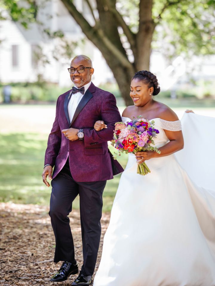 Bride and her father in purple and carrying jewel tone florals walk down the aisle.