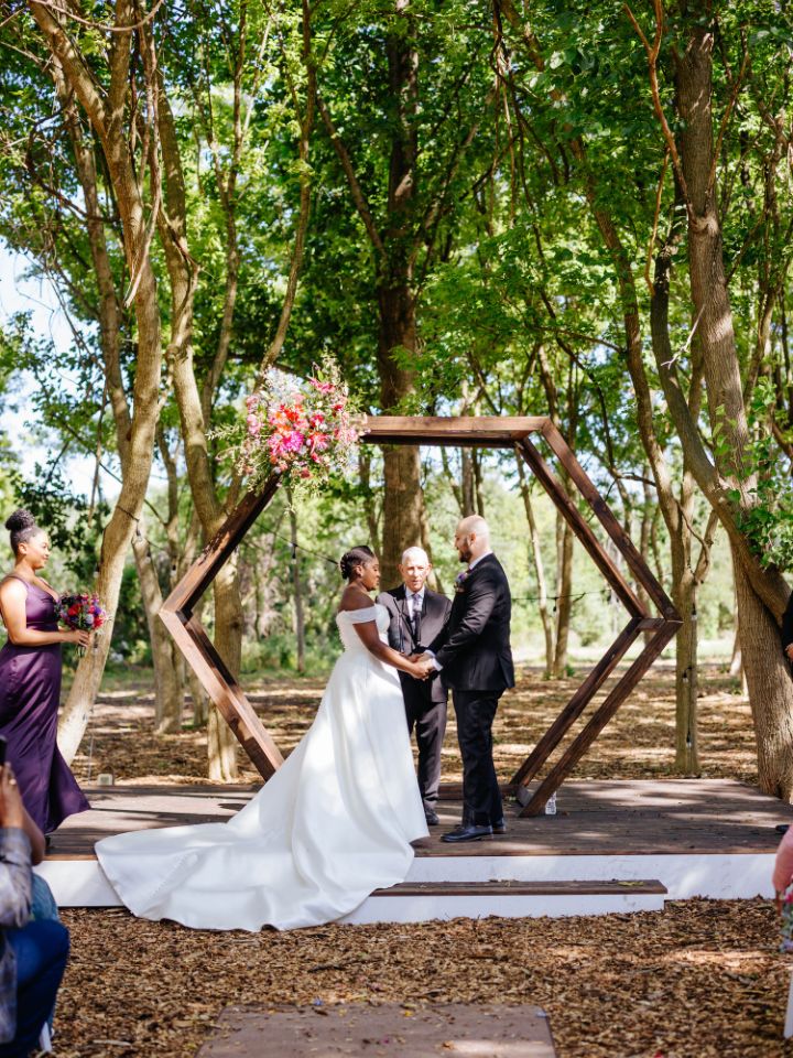 Bride and Groom stand under a hexagon arch in a wooded wedding ceremony site.