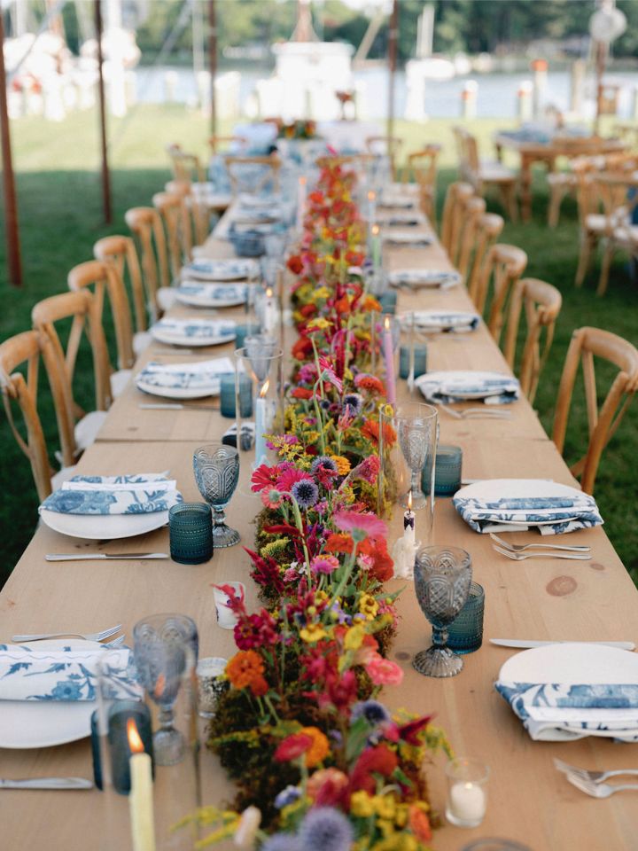 Looking down a wedding party table at an outdoor wedding reception, made up of three tables placed end to end. There is a long floral arrangement with moss and bright flowers down the entire center.