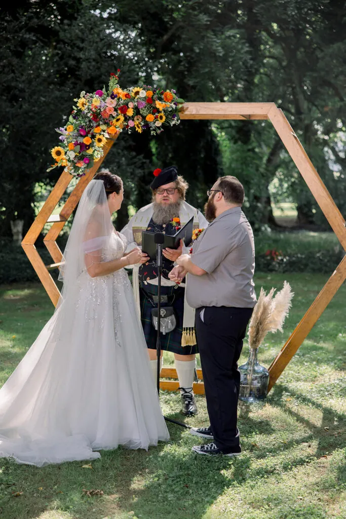 Bride and Groom say their vows under a hexagon arch covered in bright summer wedding flowers.