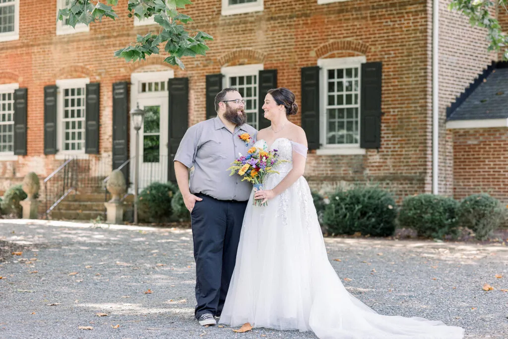 Bride and Groom smile at each other with the brick Worsell Manor in the background.